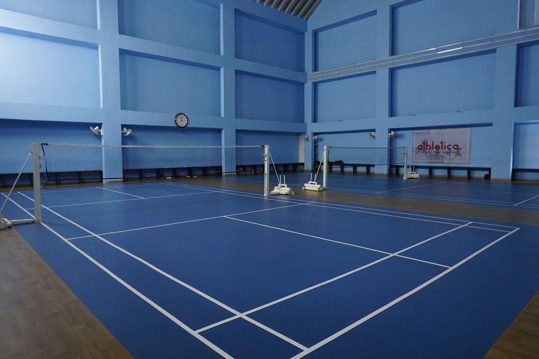 A professional-grade indoor sports hall featuring blue high-grip flooring and multiple marked badminton courts. The high ceilings and bright, even lighting create a perfect environment for competitive play and athletic training.