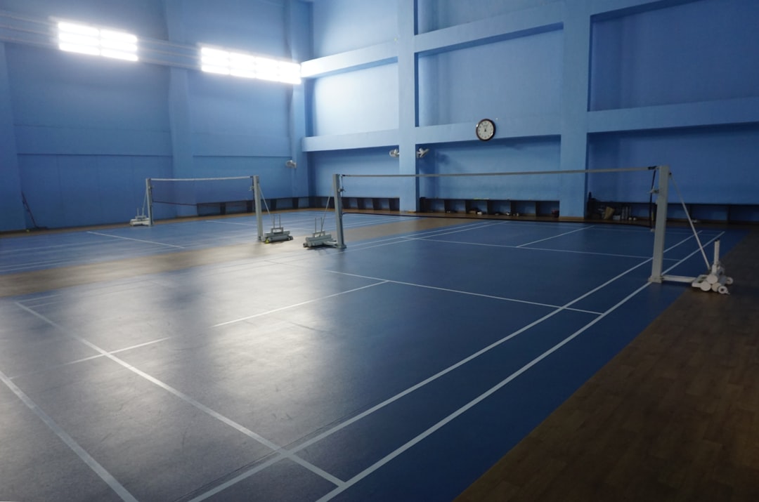 A professional-grade indoor sports hall featuring blue high-grip flooring and multiple marked badminton courts. The high ceilings and bright, even lighting create a perfect environment for competitive play and athletic training.